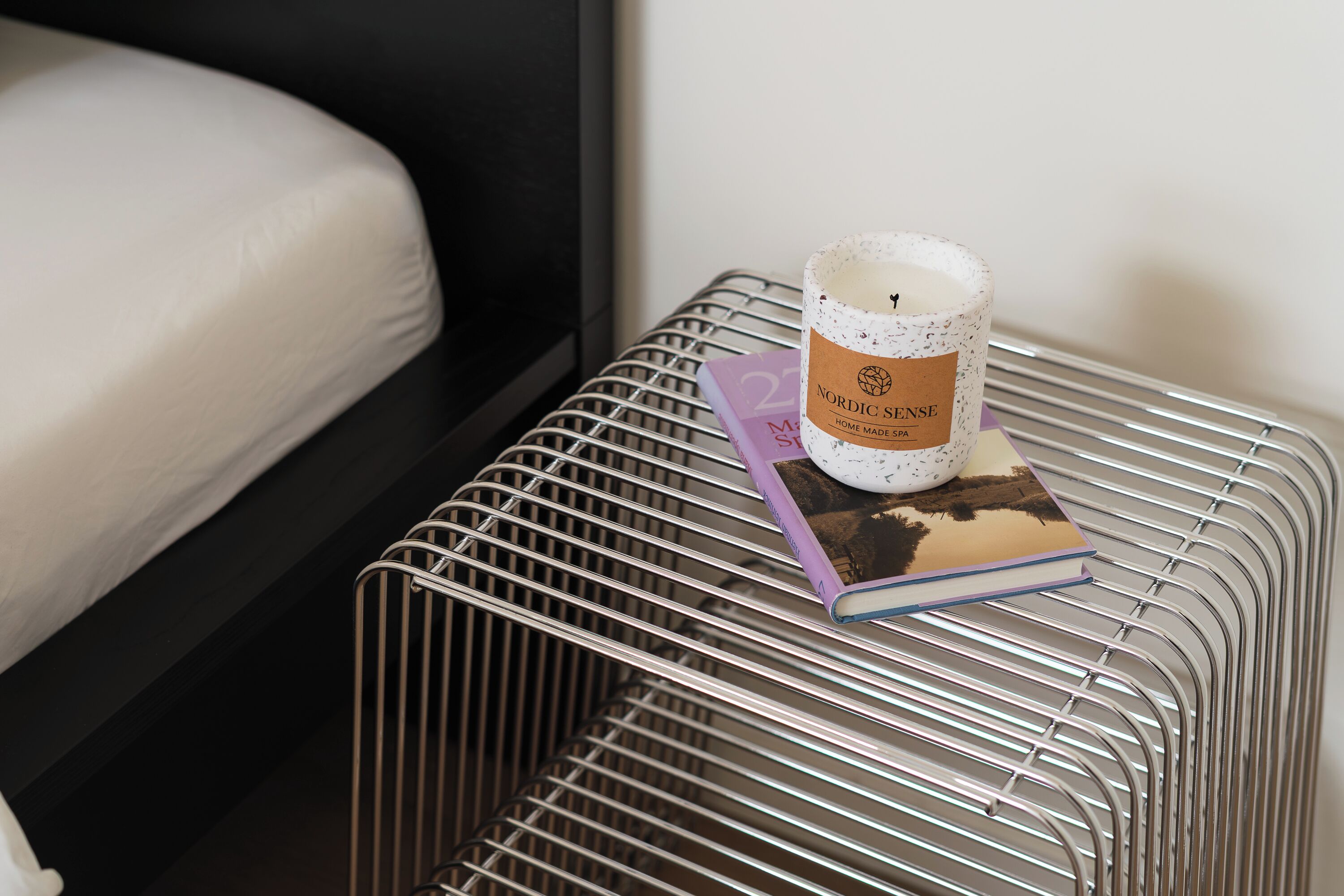 A metal wire shelf with a candle, a book, and a pillow on a bed.
