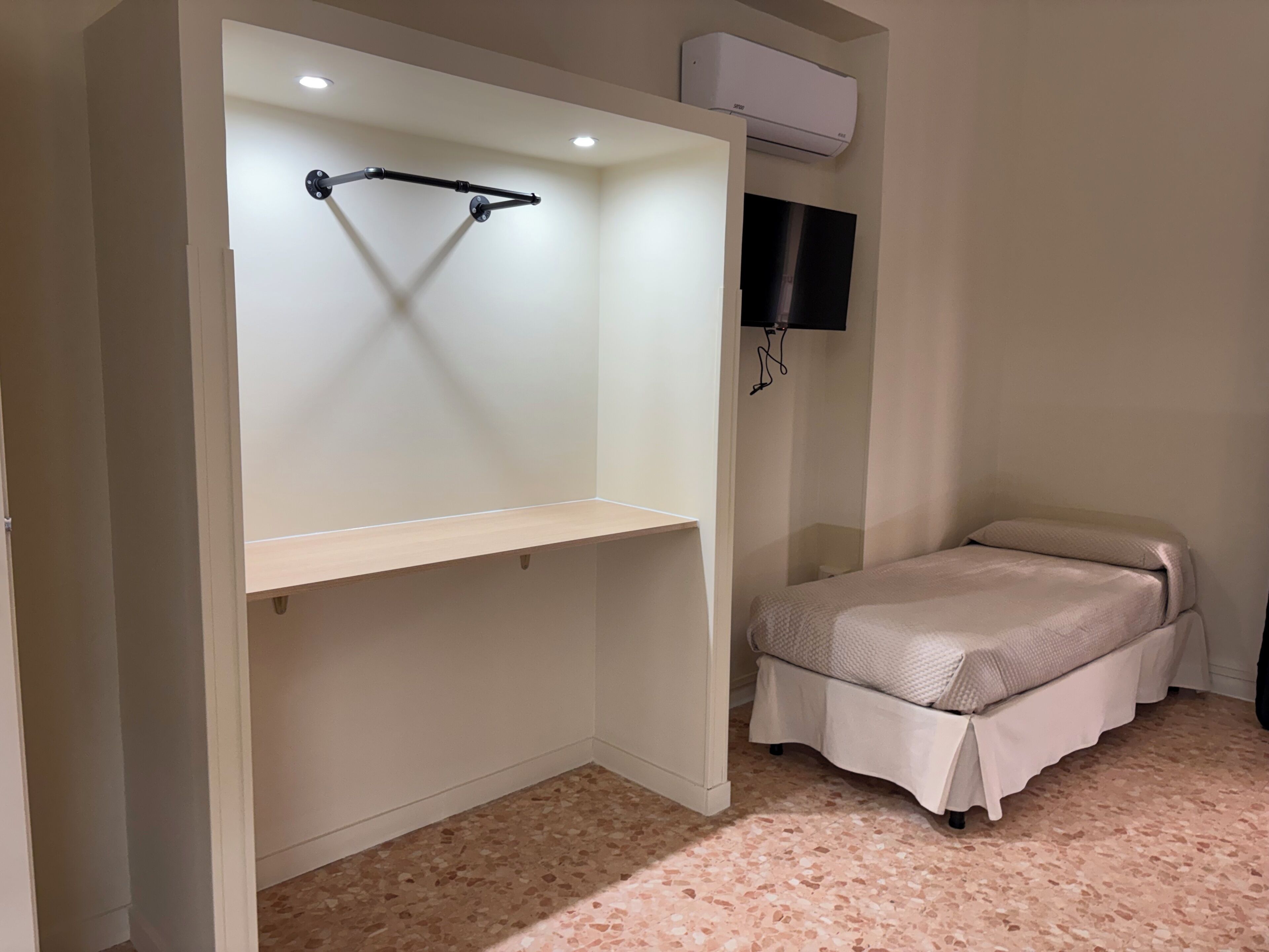 A bedroom with a bed, a built-in wardrobe, a television mounted on the wall, and an air conditioning unit above the wardrobe.