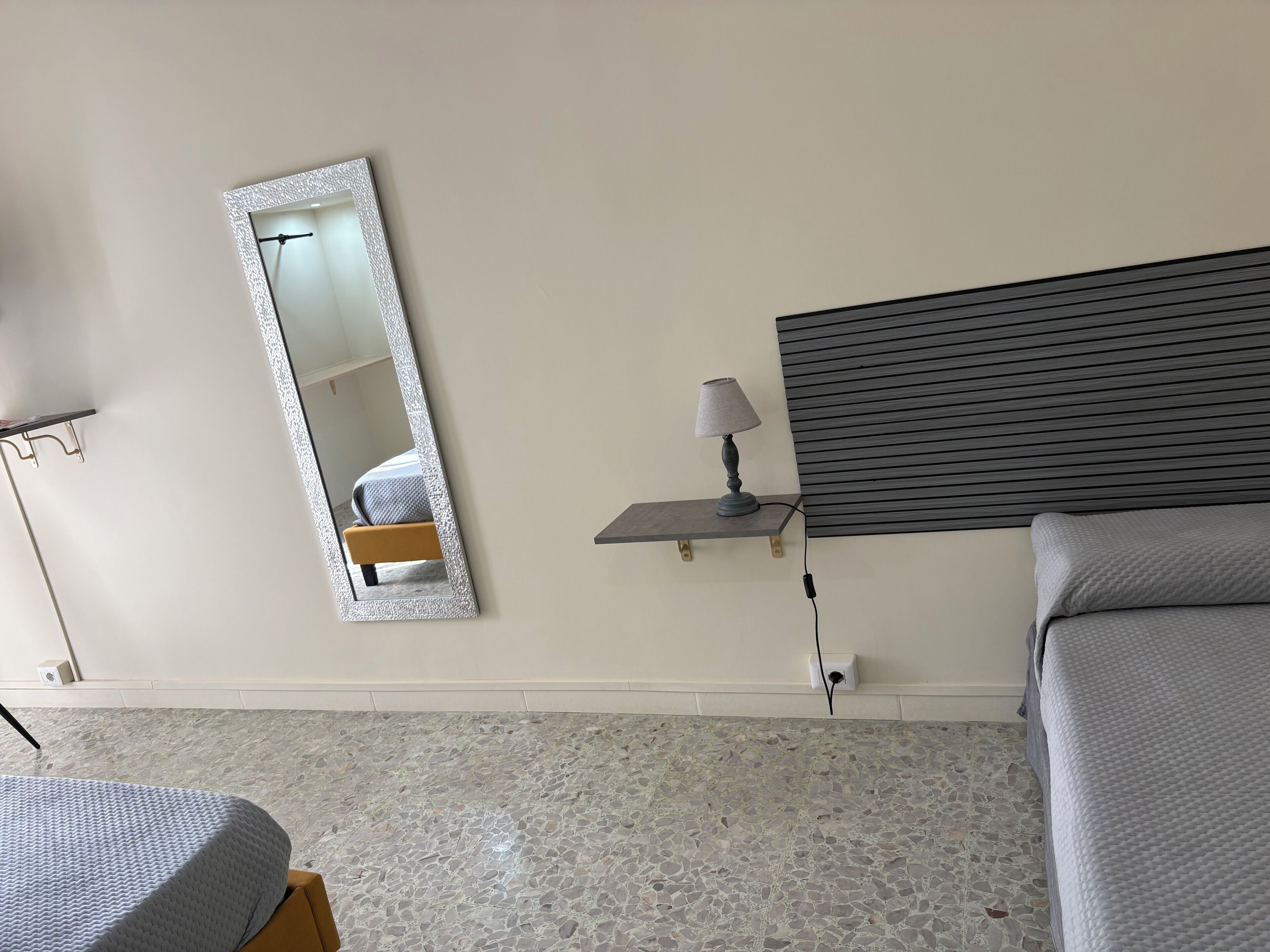 A bedroom with a bed, a bedside table with a lamp, a mirror, and a wall-mounted shelf.