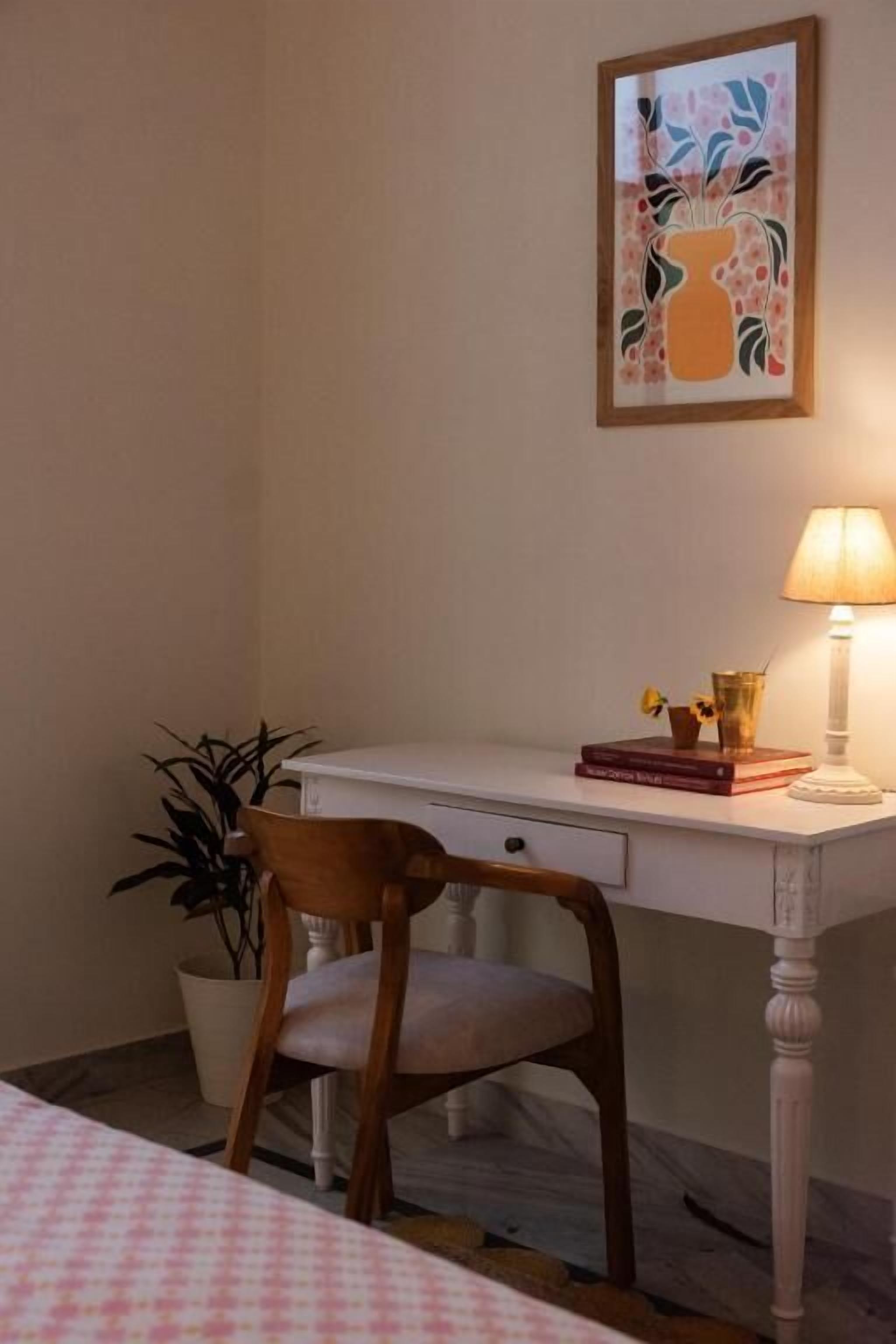 A room with a white desk, a chair, a bedside table with a lamp, a framed picture on the wall, and a potted plant.