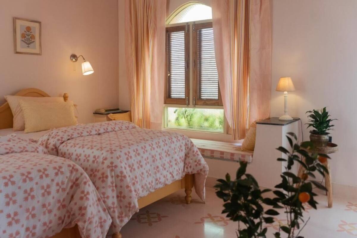 A bedroom with two beds, a window with shutters, a bedside table with a lamp, a small bench, and a potted plant.