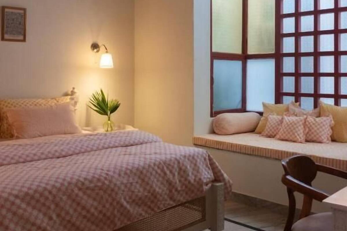 A bedroom with a bed, bedside table, lamp, window with blinds, and a chair.