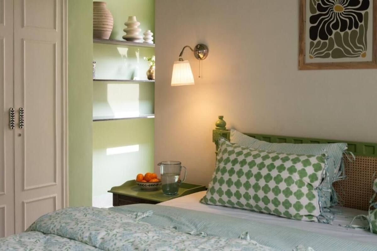 A bedroom with a bed, a nightstand, a wall-mounted lamp, and a shelf with decorative items.