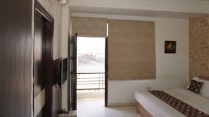 A hotel room with a bed, a television, a ceiling fan, a window with a view, and a door. - Hotel Sun Heritage (Jaipur)