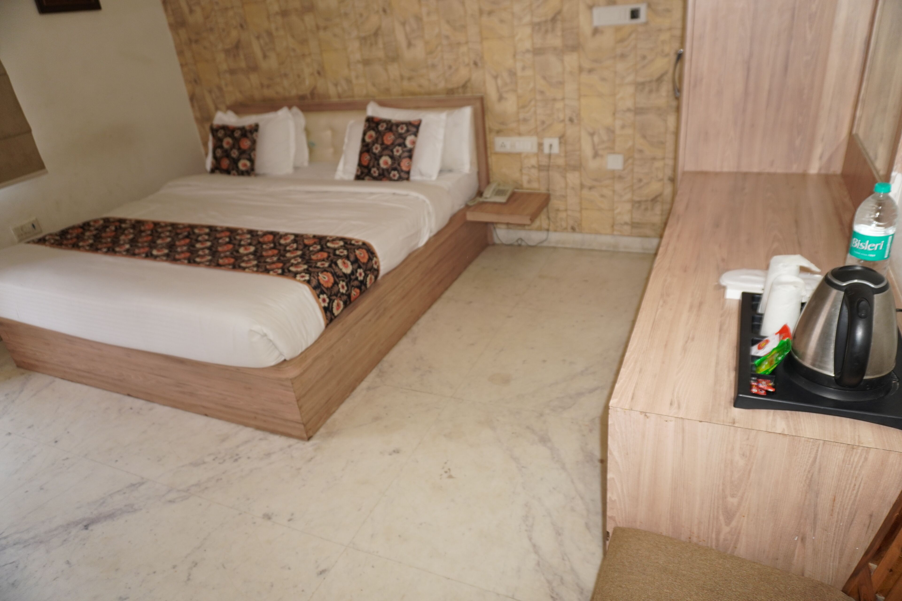 A hotel room with a bed, a wooden desk, a bottle of Bisleri water, a kettle, and a tray with a teapot.