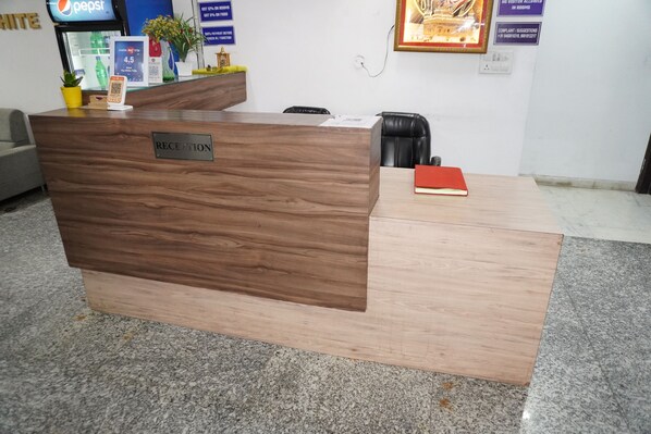 A reception desk with a wooden finish, a small potted plant, a red book, and a Pepsi refrigerator in the background.
