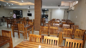 A dining area with wooden tables and chairs, a ceiling fan, and a kitchen in the background. - Hotel Sun Heritage (Jaipur)
