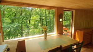 A wooden cabin with a dining table and chairs, a vase with flowers, and a view of a forest through large windows.