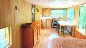 A wooden cabin interior with a bed, a dining table, a kitchen area, and a window with a view of greenery.