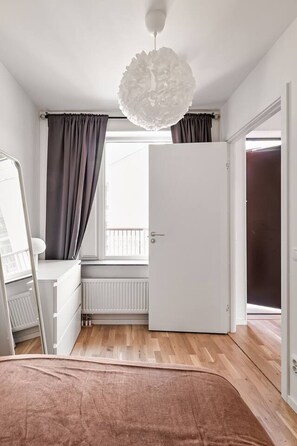 Ein Schlafzimmer mit Bett, Schminktisch, Heizkörper, einem Fenster mit Vorhängen und einer Tür.