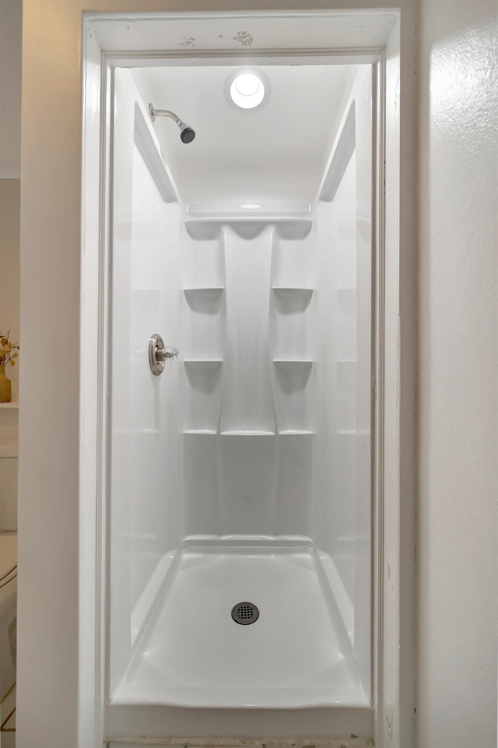 A white, enclosed shower stall with a handheld showerhead, a drain, and a small shelf.