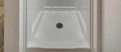 A white, enclosed shower stall with a handheld showerhead, a drain, and a small shelf.