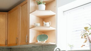 Una cocina con armarios de madera, encimera blanca, hervidor verde, pileta de acero inoxidable y una ventana con una planta.