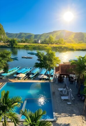 Un espace piscine avec des chaises longues et un petit pavillon, le tout entouré d’une rivière et de collines.