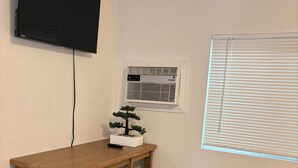 A room with a wooden table, two chairs, a television mounted on the wall, an air conditioning unit, and a window with blinds.