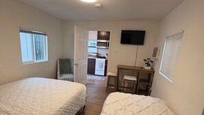 A bedroom with two beds, a TV, a small table, and a kitchenette in the background.