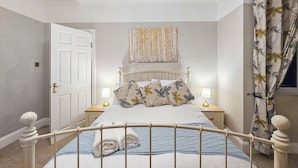 Un lit bien fait, avec une tête de lit en métal, deux tables de chevet avec des lampes, une porte et un rideau à motifs floraux.