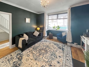 A living room with a dark green wall, a patterned rug, a black sofa, a blue armchair, a small side table with a lamp, and a window with blinds.