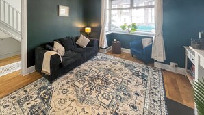 Un salon avec un mur vert foncé, un tapis à motifs, un canapé noir, un fauteuil bleu, une petite table d’appoint avec une lampe et une fenêtre avec des stores.