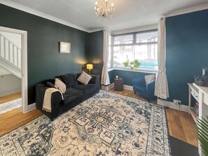 A living room with a dark green wall, a patterned rug, a black sofa, a blue armchair, a small side table with a lamp, and a window with blinds. - Gorgeous house in a great location for exploring East Kent at its finest. (Ramsgate)