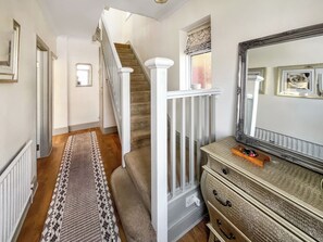 A hallway with a patterned rug, a staircase with white railings, a dresser with a mirror, and a radiator.
