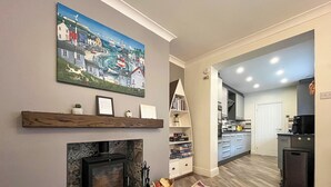 Uma sala de estar com lareira, um tapete colorido, uma estante de livros e um quadro com uma paisagem costeira.