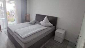 A modern bedroom with a large bed, a bedside table, a nightstand, and a view through a glass door.
