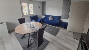 A modern living room with a round dining table, two sofas, a small kitchen area, and a window with a view of buildings.