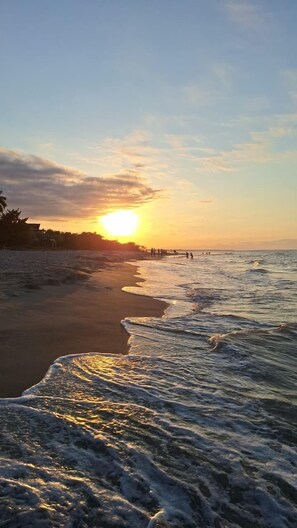Ein Strand mit Wellen, einem Sonnenuntergang und einer fernen Küstenlinie mit Gebäuden.