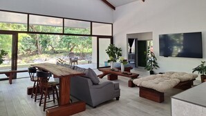 Amplia sala de estar con un ventanal grande, vigas de techo de madera, un sofá gris, una mesa de centro de madera y un televisor montado en la pared.