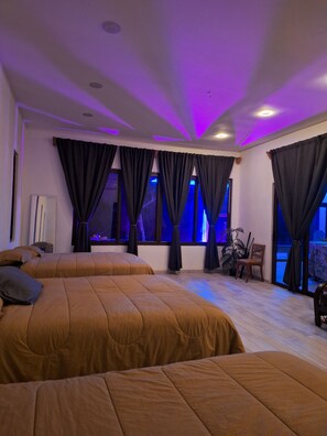 A hotel room with two beds, large windows with curtains, a plant, and a chair.