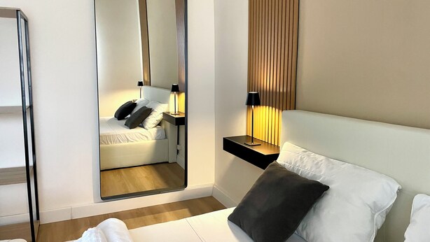 A modern hotel room with a bed, a bedside table with a lamp, a mirror, and a window with blinds. - Rome Vatican/ Dimora 63 AIR CONDITIONING & WI-FI Premium Apartment (Roma)