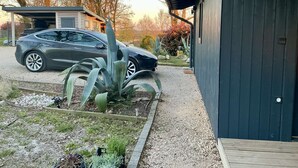 A residential area with a car parked in front of a house, a garden with various plants, and a wooden deck.