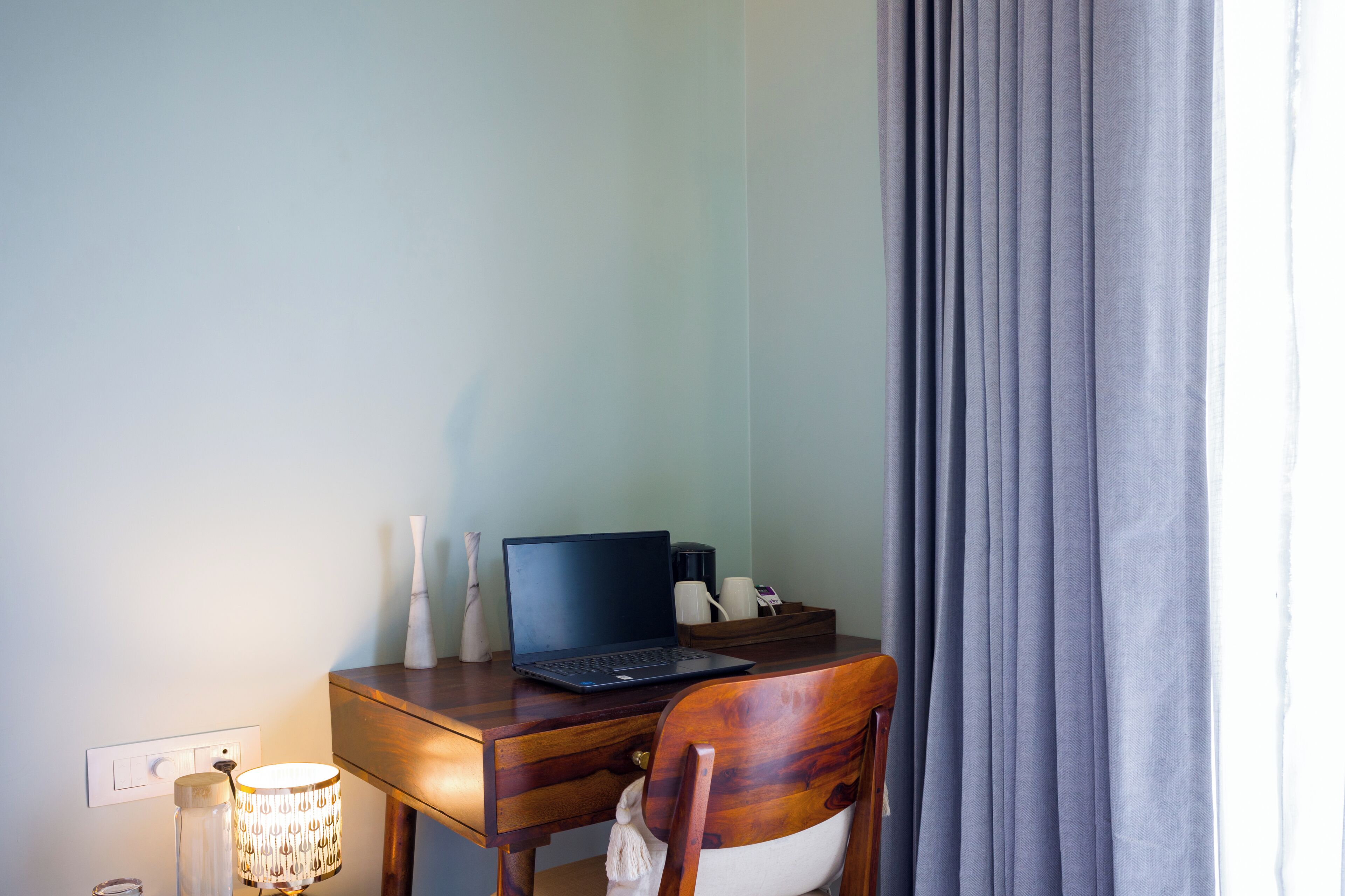 Et træskrivebord med en bærbar computer, en lampe og en stol i et rum med lyse vægge og en grå gardin.