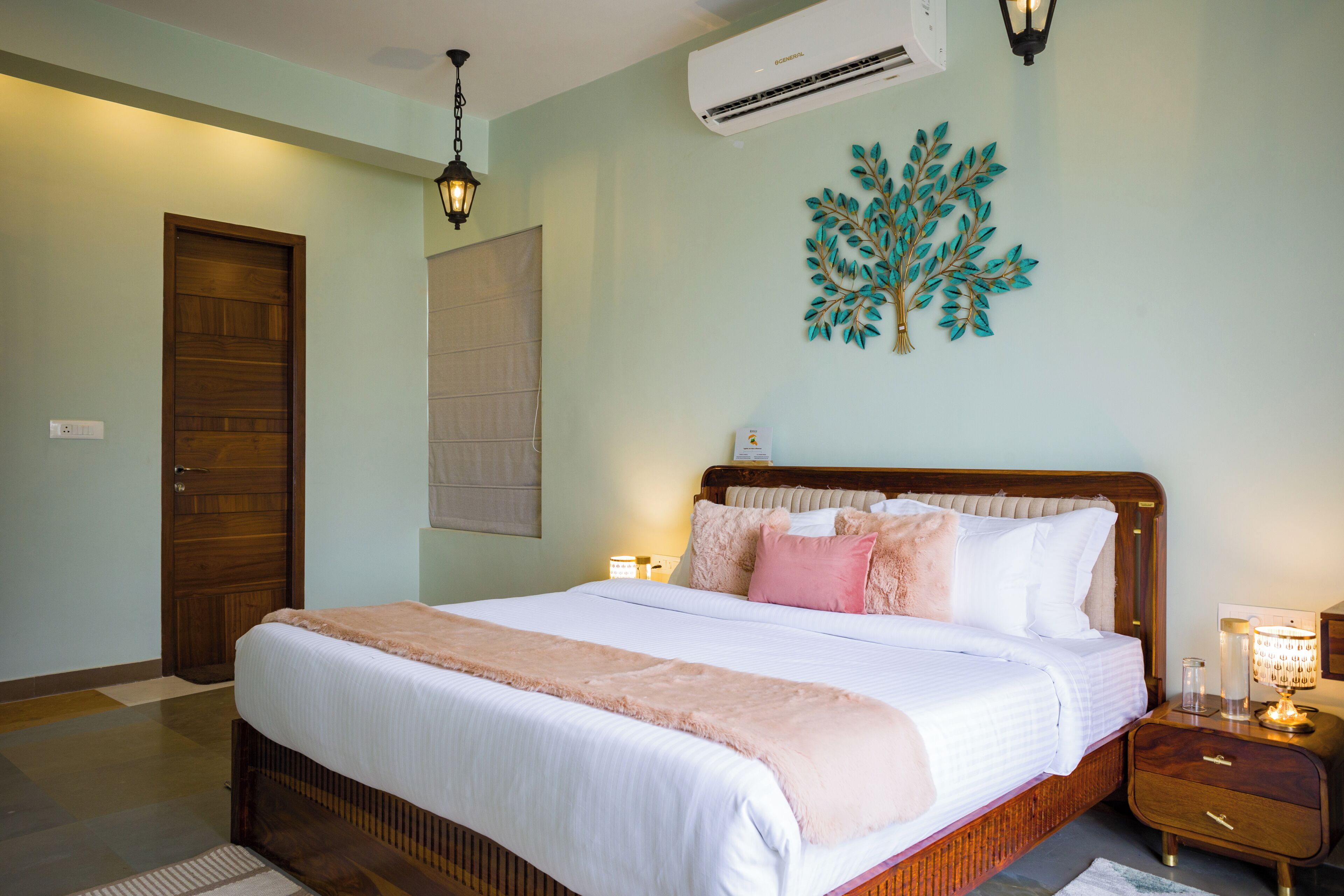 A neatly made bed with a wooden headboard, a wall-mounted air conditioner, a decorative tree, and a nightstand with a lamp.