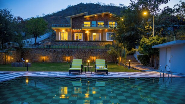 Una piscina con baldosas a cuadros, dos sillas de descanso y una casa con balcón.