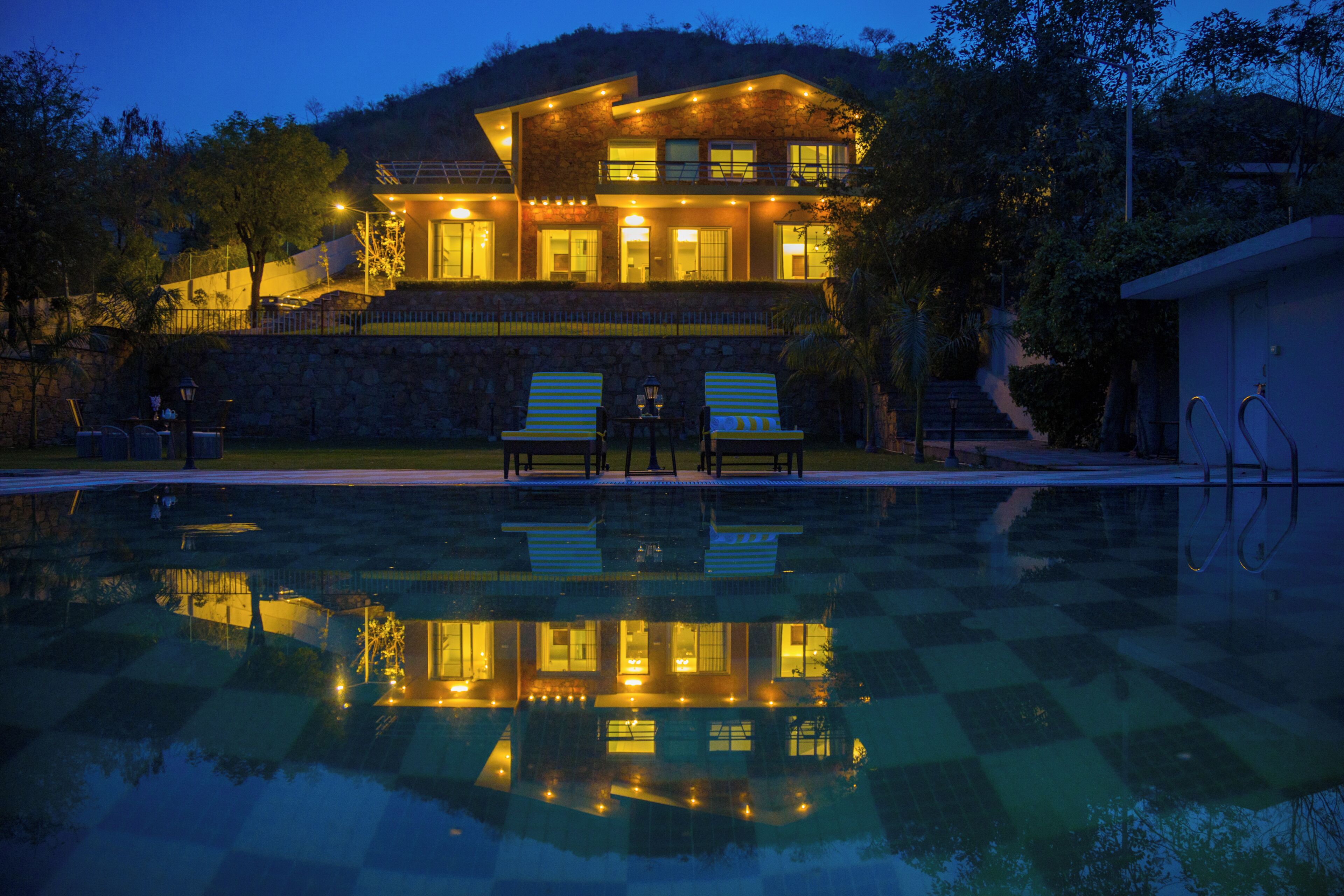 A two-story house with a swimming pool in front, stone walls, and a balcony with lights.
