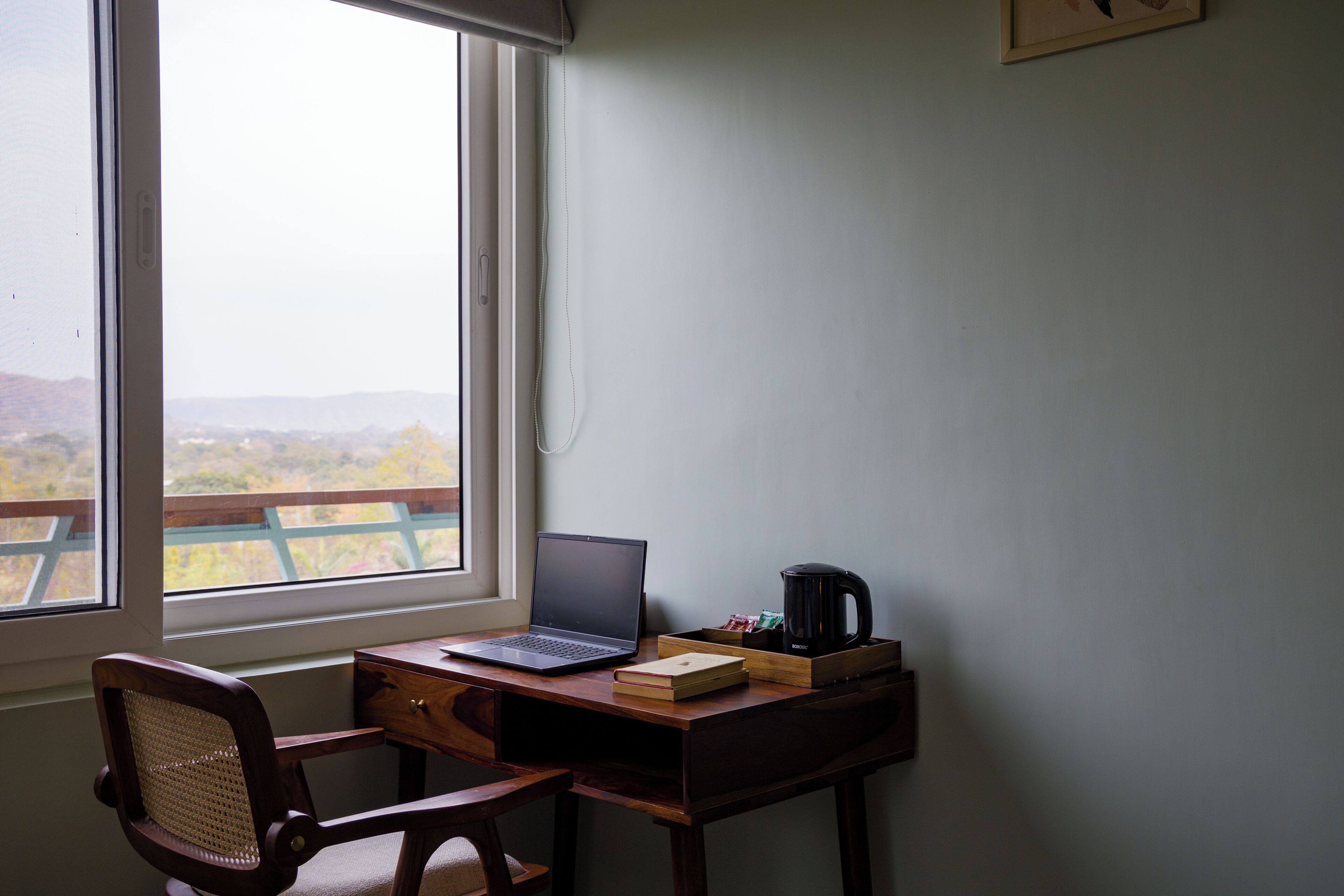 A room with a desk, laptop, coffee maker, and a view of a balcony with a railing.