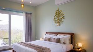 Un dormitorio con una cama grande, una mesita de noche con lámpara, un adorno de árbol en la pared y una ventana con cortinas.