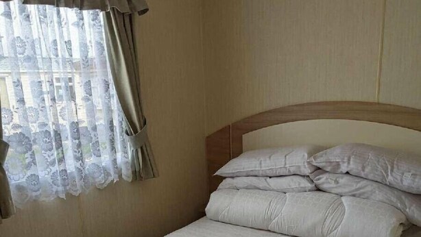 Cama bem arrumada com cabeceira de madeira em quarto com paredes bege e janela com cortinas leves.