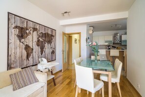 Een eethoek met een glazen tafel, witte stoelen, een groot wereldkaart schilderij, en een keuken op de achtergrond.