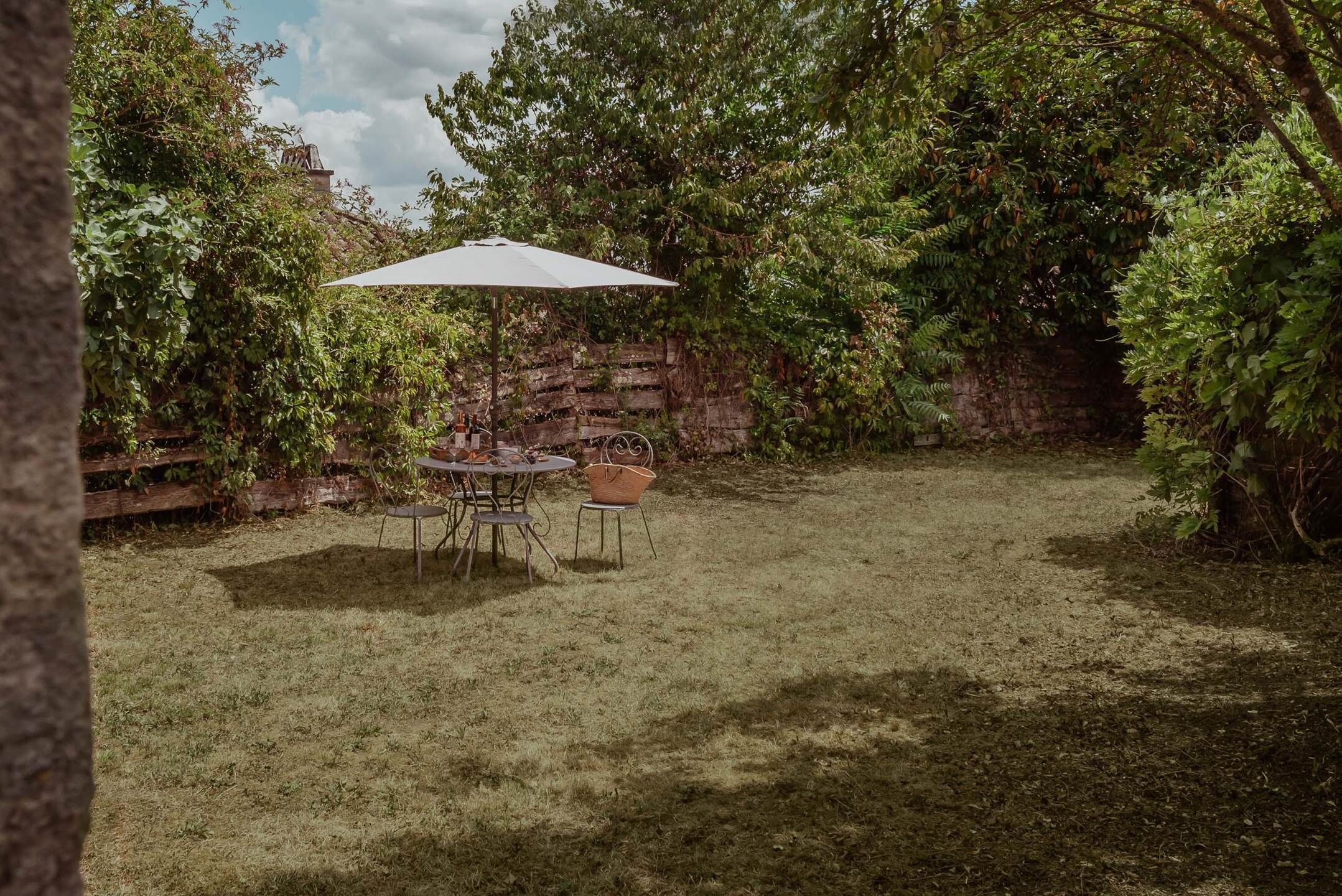 A patio area with a table and chairs under a white umbrella, surrounded by dense foliage and a brick wall.