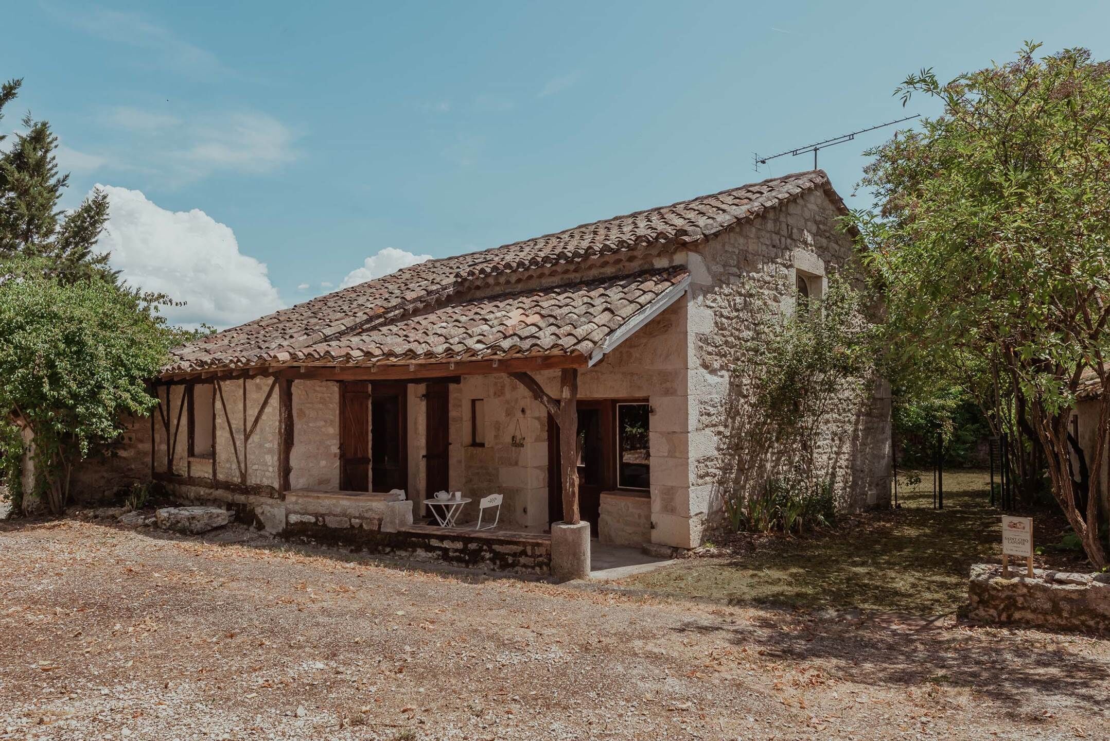 A stone building with a tiled roof, a porch with chairs, and a gravel ground.