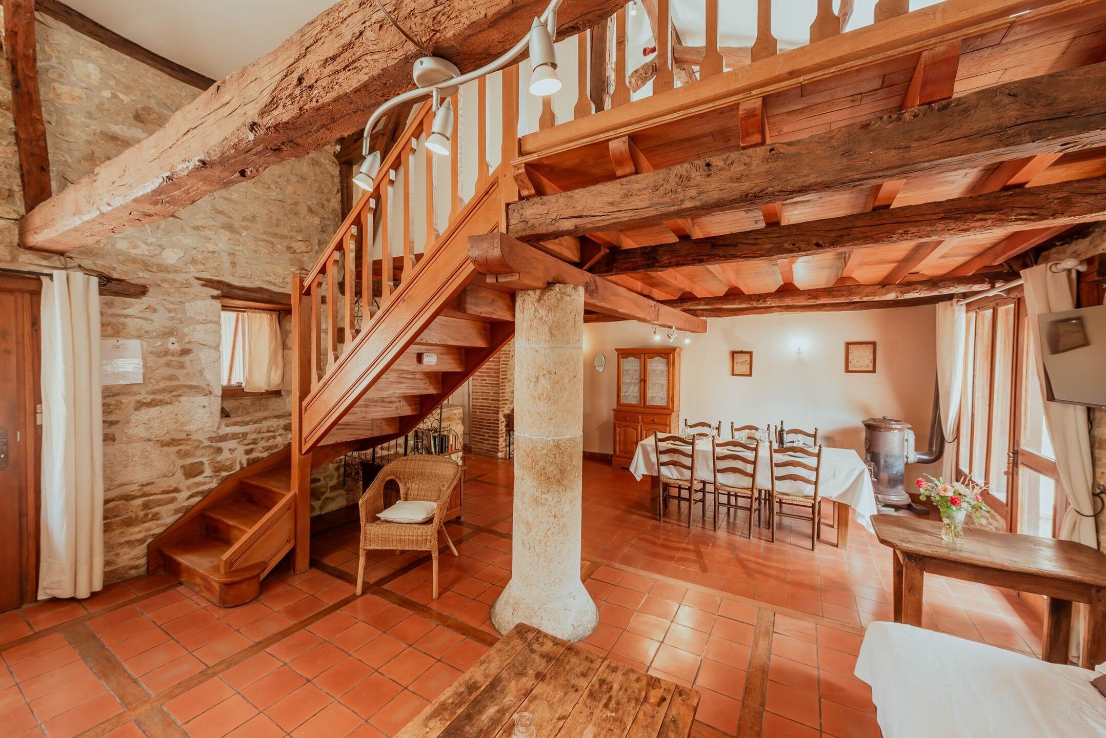 A rustic interior with exposed wooden beams, a stone wall, a wooden staircase, a dining area with chairs and a table, and a large pillar.