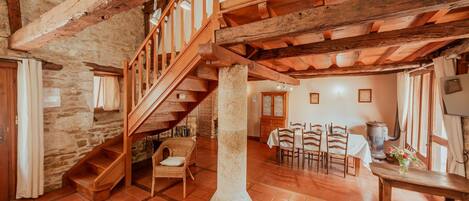 A rustic interior with exposed wooden beams, a stone wall, a wooden staircase, a dining area with chairs and a table, and a large pillar.