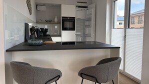 A modern kitchen with a central island, two bar stools, and a large window with blinds. - Strandwiese - Baltic Sea Happiness - "Baltic Sea Happiness"... The sound of the sea included! (Dahme)
