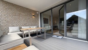 A modern outdoor living space with a brick wall, a wooden deck, a white sofa, and a glass door leading to an interior dining area. - Strandwiese - Baltic Sea Happiness - "Baltic Sea Happiness"... The sound of the sea included! (Dahme)