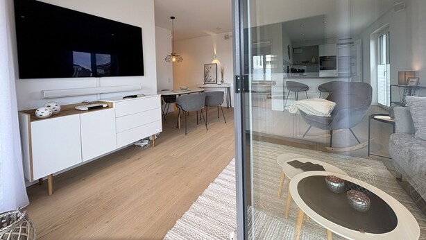 A modern living room with a large flat-screen TV, a dining area with chairs, and a kitchen in the background. - Strandwiese - Baltic Sea Happiness - "Baltic Sea Happiness"... The sound of the sea included! (Dahme)