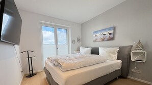 A bedroom with a bed, a television, a window, and a painting on the wall. - Strandwiese - Baltic Sea Happiness - "Baltic Sea Happiness"... The sound of the sea included! (Dahme)