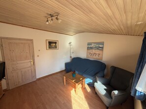 A room with a wooden ceiling, a door, a blue couch, a small table with a plant, and two wall-mounted pictures. - Apartment 3 Geltinger Bucht - relaxation near the beach & nature (Gelting)